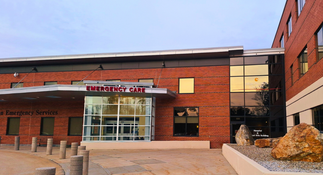 Monadnock Community Hospital emergency department entrance. The building features brick walls and large glass windows, with signage reading 'Emergency Care' above the entrance