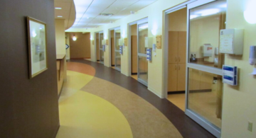 A modern and clean hallway in the emergency department of Monadnock Community Hospital, featuring several private patient rooms with glass doors and well-lit, curved pathways. Alt text for second photo: The reception and nurse's station area in the emergency department of Monadnock Community Hospital, showcasing a well-organized workspace with office equipment, floral arrangements, and a view of patient rooms in the background.