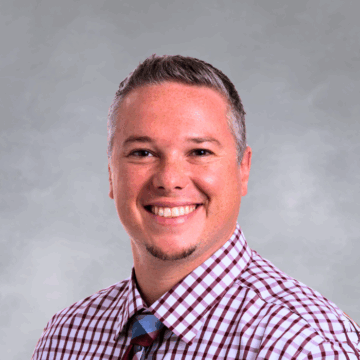 Smiling man with short gray hair wearing a red and white checkered shirt and a multicolored plaid tie, in front of a soft gray studio background. Ask ChatGPT 