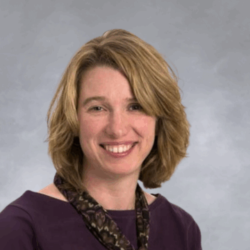 Smiling woman with short, layered blonde hair wearing a dark plum top and a patterned scarf, photographed against a white background.