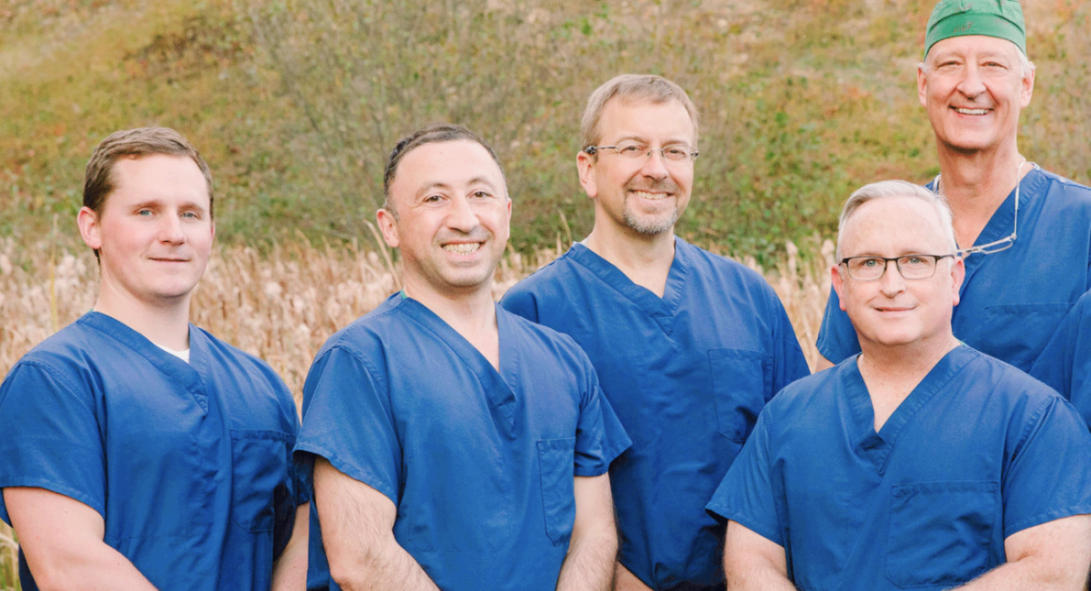 A group photo of five male orthopedic surgeons from Monadnock Orthopedic Associates standing outside in front of a natural backdrop. They are all wearing blue surgical scrubs and are smiling at the camera by Brianna Morrissey ©Monadnock Studios by BLM Photography 2022