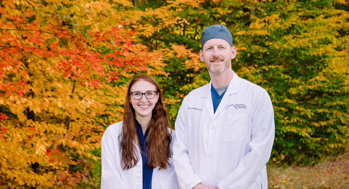 A photo of two orthopedic specialists from Monadnock Orthopedic Associates standing outside in front of autumn foliage. The female specialist on the left is wearing glasses and a white coat, and the male specialist on the right is wearing a white coat and a surgical cap. Both are smiling at the camera by Brianna Morrissey ©Monadnock Studios by BLM Photography 2022