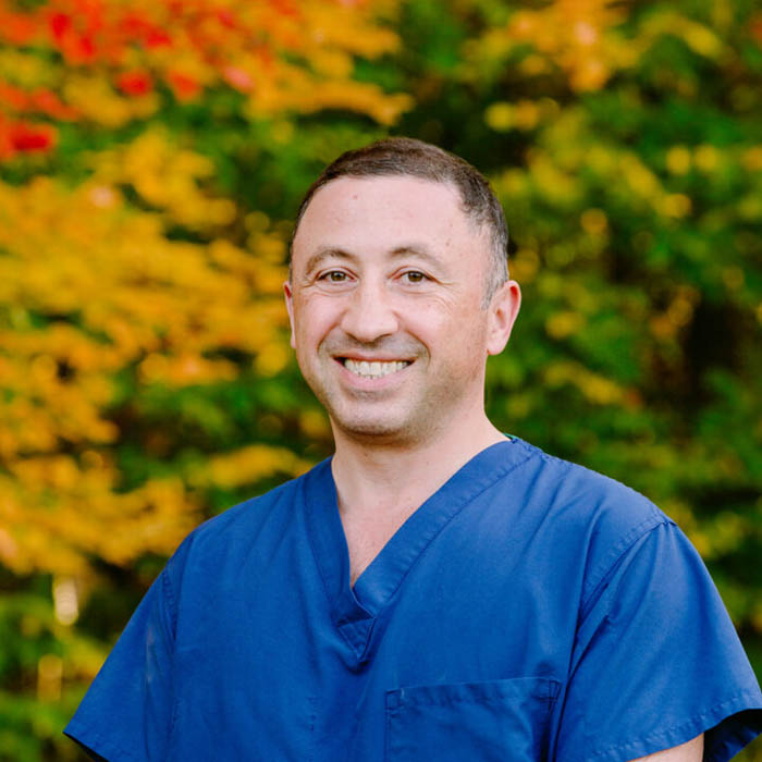 Vache Hambardzumyan wearing blue scrubs stands smiling against a backdrop of vibrant autumn foliage with green, yellow, and red leaves. He has short dark hair and appears to be outdoors