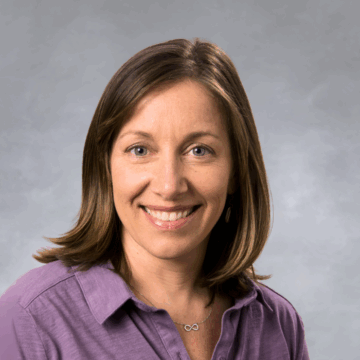 Smiling woman with shoulder-length light brown hair wearing a lavender button-down shirt and an infinity necklace, in front of a soft gray studio background.