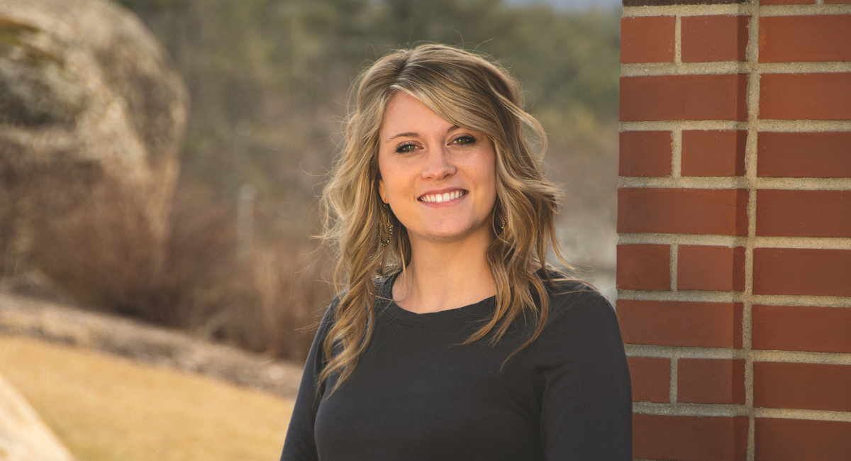 A smiling woman with blonde hair standing outside near a brick wall, with a backdrop of natural scenery, reflecting a friendly and approachable demeanor.