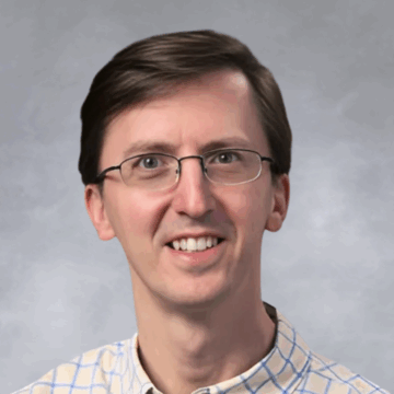Smiling man with short brown hair and glasses, wearing a light blue plaid button-down shirt, in front of a soft gray studio background.