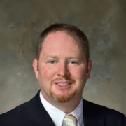 Smiling man with short light brown hair and a goatee, wearing a black suit, white shirt, and yellow striped tie, posed in front of a mottled brown-gray background.