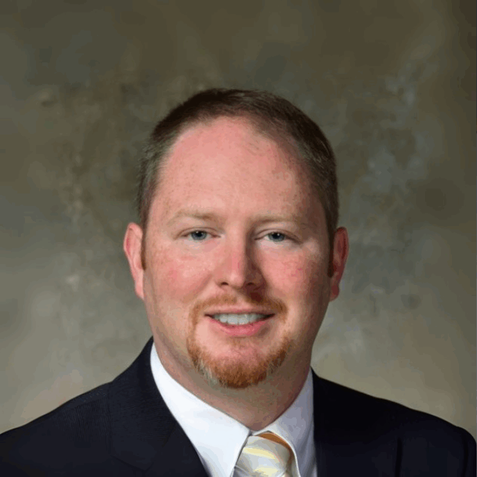 Smiling man with short light brown hair and a goatee, wearing a black suit, white shirt, and yellow striped tie, posed in front of a mottled brown-gray background.