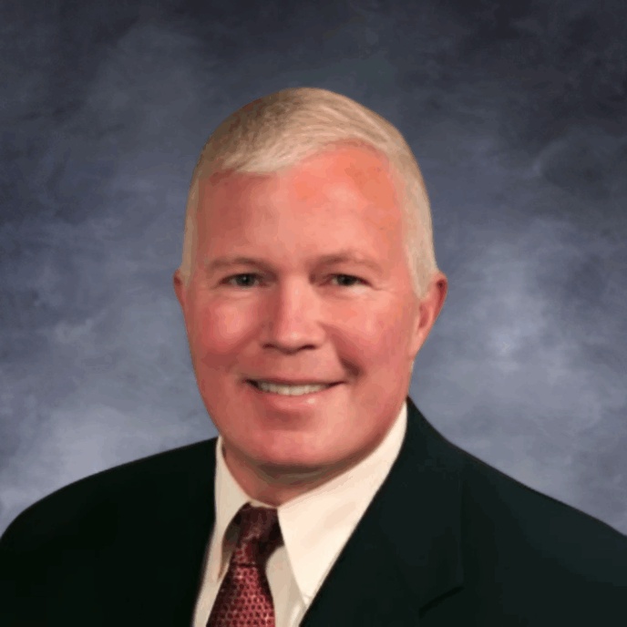 A man with short gray hair is smiling at the camera. He is wearing a dark suit jacket, a white dress shirt, and a red patterned tie. The background is softly blurred in warm neutral tones.