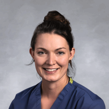 Smiling woman with dark brown hair tied up in a bun, wearing navy blue medical scrubs and gold earrings, in front of a soft gray studio background.