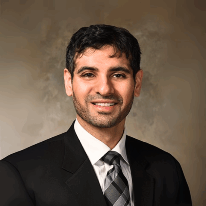 Smiling male clinician with short dark hair and trimmed beard, wearing a black suit and striped tie, posed against a neutral background.
