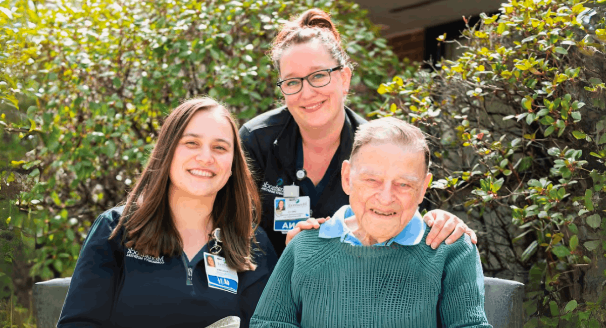 David Lang, seated and smiling, is flanked by two caring MCH staff members, Emily Shaw (CNA) and Katelynn Kelleher (RN), in an outdoor setting at Monadnock Community Hospital.
