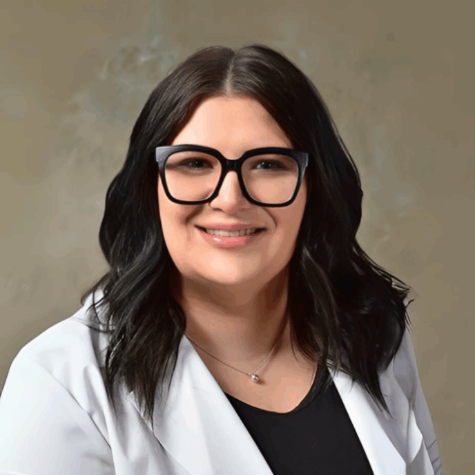 Professional headshot of a woman with long dark hair, wearing black glasses, a white coat, and a black top, smiling against a neutral backdrop
