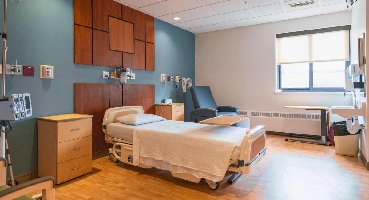 Private hospital room with a patient bed, medical equipment, natural light from a window, and a comfortable chair for recovery.