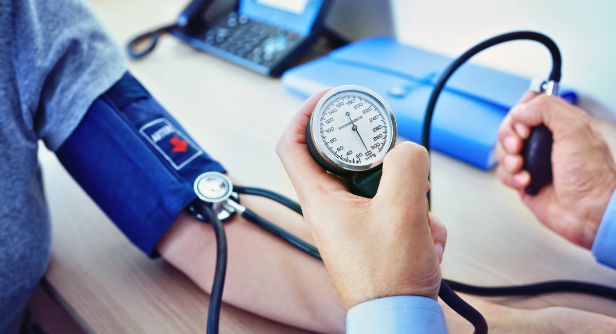 A healthcare professional measures a patient's blood pressure using a sphygmomanometer. The patient's arm is wrapped in a blue blood pressure cuff and the healthcare professional is holding the gauge and bulb monitoring the readings. A stethoscope is visible around the professional's neck and there is a laptop on the desk in the background