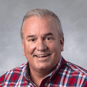 Smiling man with short gray hair wearing a red, white, and blue plaid button-down shirt, in front of a soft gray studio background.