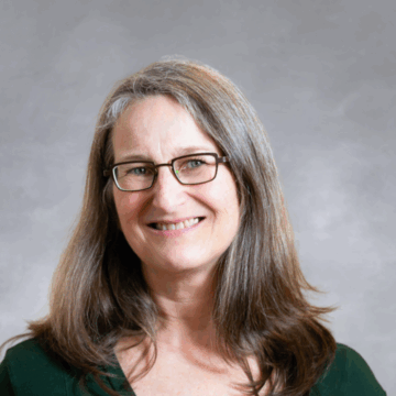 Smiling woman with long, straight gray hair and glasses, wearing a dark green top, in front of a soft gray studio background.