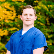 Serious-looking man with short light brown hair wearing blue scrubs, standing outside in front of green and yellow fall leaves.
