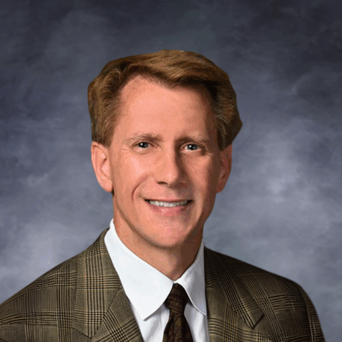 Professional portrait of a middle-aged man with light skin and neatly styled light brown hair, wearing a brown plaid suit jacket, white dress shirt, and dark patterned tie, smiling in front of a studio-style dark gray background.