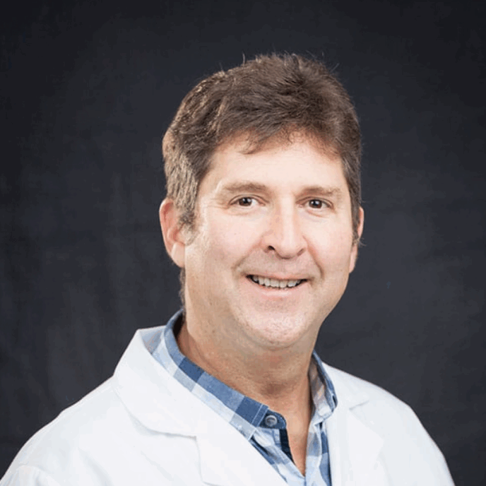 Smiling male clinician with short brown hair, wearing a white medical coat over a blue plaid shirt, posed against a dark background.