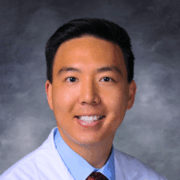Smiling man with short black hair, wearing a white medical coat, light blue shirt, and red patterned tie, posed in front of a soft gray studio background.