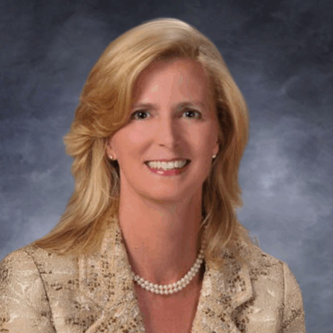 A woman with shoulder-length blonde hair is smiling at the camera. She is wearing a light gold patterned jacket, a pearl necklace, and small earrings. The background is a neutral cloudy gradient in shades of gray and blue.