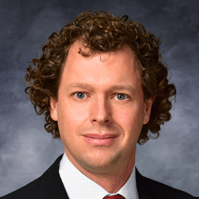 Smiling man with fair skin and curly light brown hair, wearing a black suit jacket, white shirt, and red tie, posed in front of a soft gray studio background.