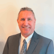 Man with short gray hair smiling, wearing a gray suit, white shirt, and patterned tie, in front of a light background.