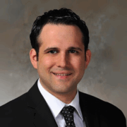 Professional headshot of a man with short dark hair, wearing a black suit, white shirt, and black patterned tie, smiling against a neutral background