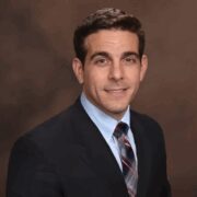 Professional headshot of a man in a dark suit, light blue shirt, and striped tie, smiling in front of a brown backdrop.
