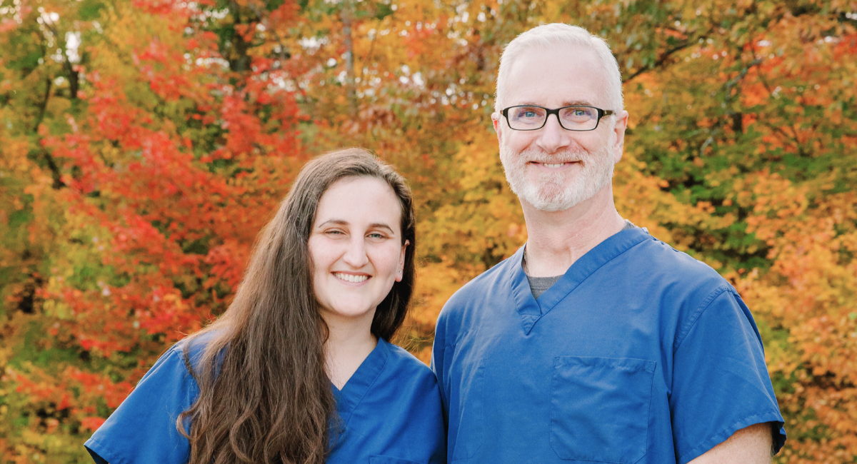 Kayleigh Sullivan and Dr Shawn P Harrington Two healthcare professionals in blue scrubs standing in front of autumn foliage smiling at the camera by Brianna Morrissey ©Monadnock Studios by BLM Photography 2022