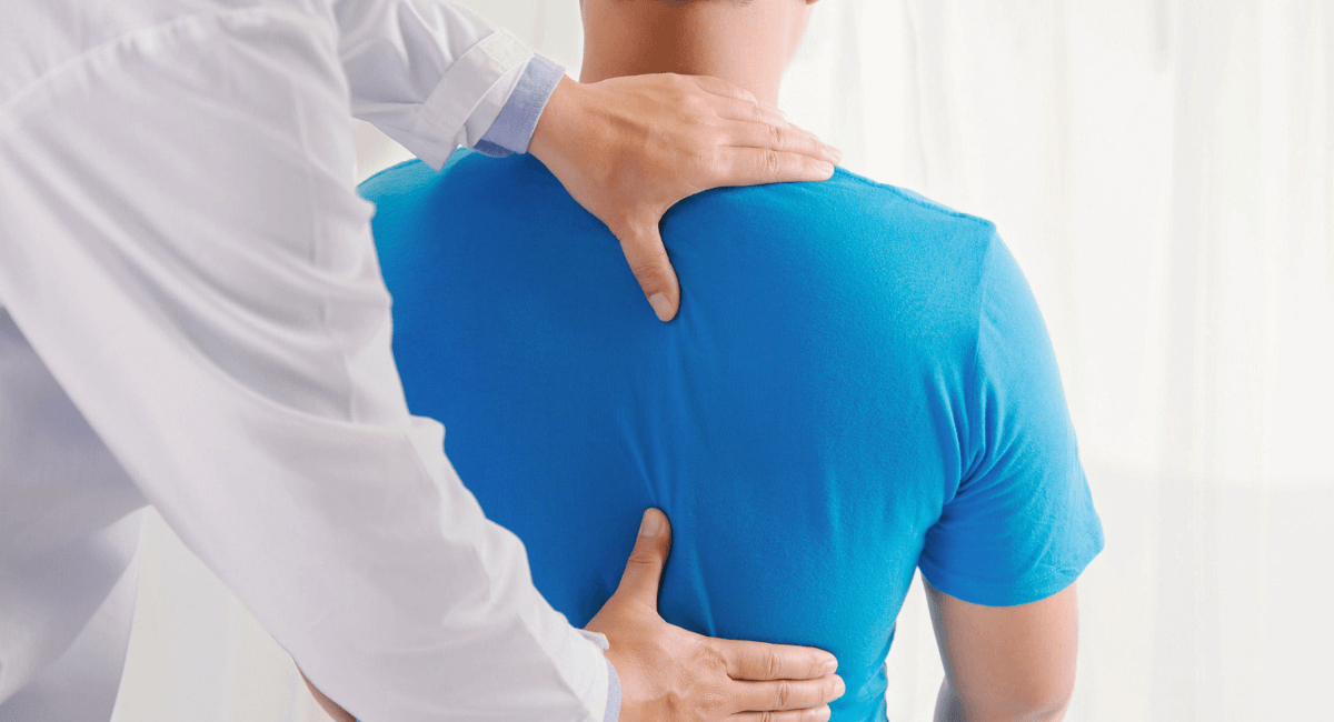 A healthcare professional examines a patient’s upper back and spine. The patient is wearing a blue shirt and is seated, while the provider uses both hands to gently assess the back area.