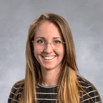 Smiling woman with long straight blonde hair and glasses, wearing a black top with white stripes, in front of a soft gray studio background.