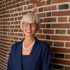 Cynthia K. McGuire, President & CEO of Monadnock Community Hospital, smiling and wearing a navy blue suit with a pearl necklace, standing against a brick wall.