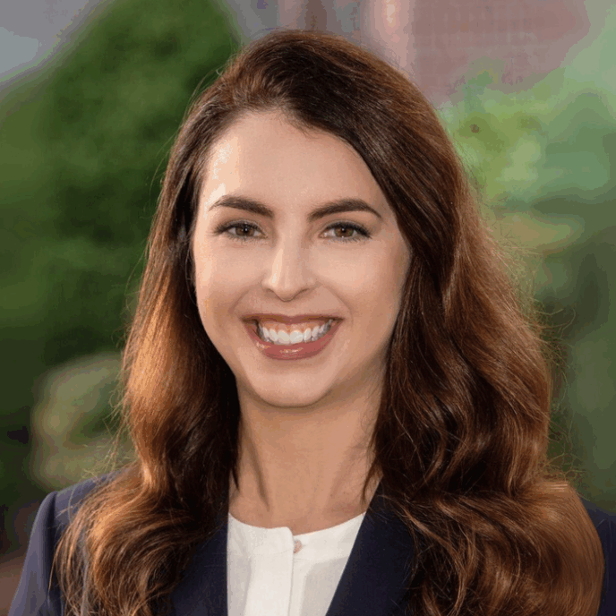A woman with long brown hair, wearing a navy blazer and white blouse, smiles outdoors with greenery and brick in the background.