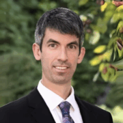 A man with short salt-and-pepper hair, wearing a black suit and patterned tie, stands outdoors with greenery in the background.