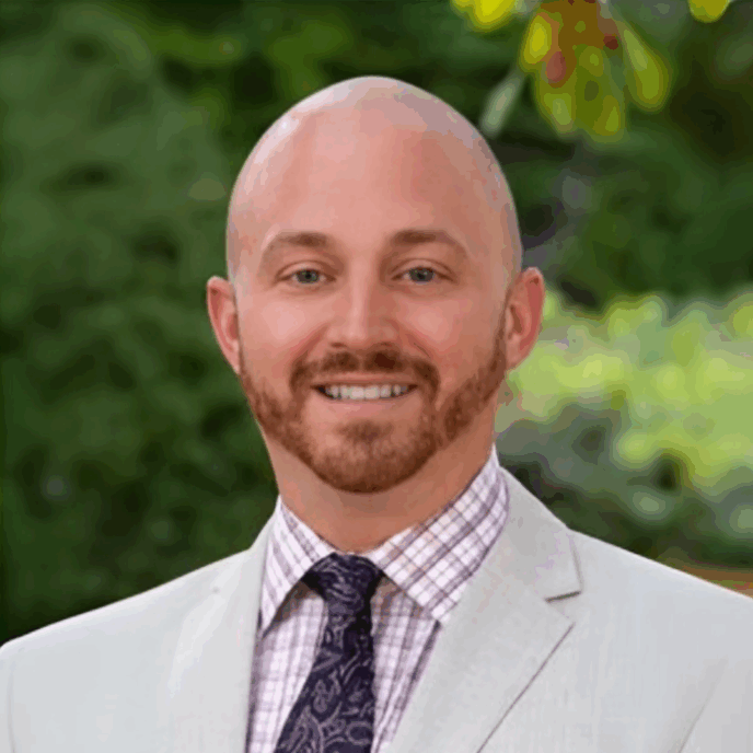 A man with a bald head and trimmed beard is smiling at the camera. He is wearing a light beige suit jacket over a checkered shirt with a dark paisley tie. The background is outdoors with green foliage.