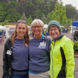 LeeAnn Clarke Moore, Cyndee McGuire, and Laura Gingras smiling and posing outdoors at a community event. They are wearing casual attire, with two wearing Monadnock Community Hospital shirts and name badges. The event tents and trees are visible in the background, indicating a lively and friendly gathering