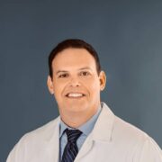 A general surgeon, stands in front of a blue backdrop wearing a white lab coat and dark tie. He is smiling with a professional, approachable expression.