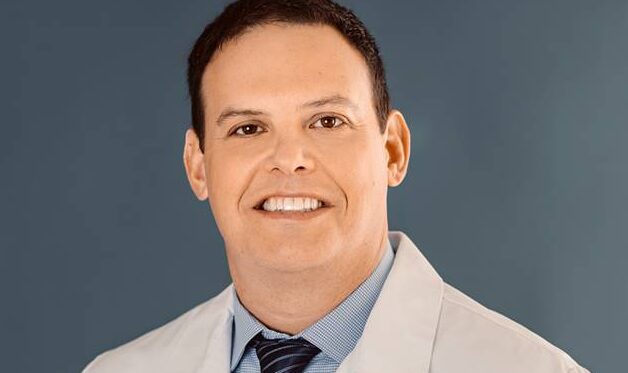 A general surgeon, stands in front of a blue backdrop wearing a white lab coat and dark tie. He is smiling with a professional, approachable expression.