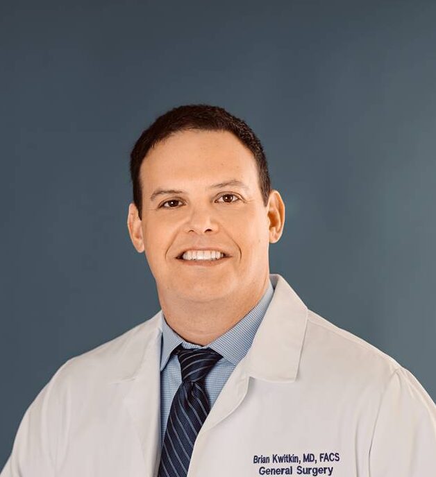 A general surgeon, stands in front of a blue backdrop wearing a white lab coat and dark tie. He is smiling with a professional, approachable expression.