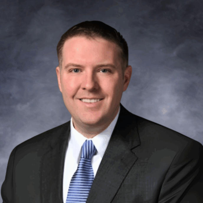Man with short brown hair smiling, wearing a dark suit, white shirt, and blue striped tie, in front of a neutral background.