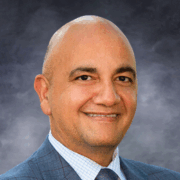 Smiling man with a bald head, wearing a blue checkered suit jacket, light blue checkered shirt, and dark tie, posed in front of a soft gray studio background.