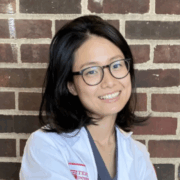 A smiling woman with shoulder-length dark hair, glasses, and a white medical coat sits in front of a brick wall.