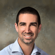 Smiling man with short dark hair and trimmed beard, wearing a light gray dress shirt, posed in front of a mottled tan background