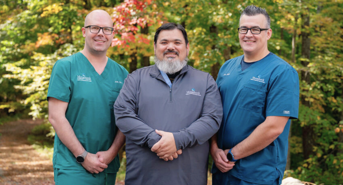 Three healthcare professionals from Monadnock Community Hospital standing outdoors in front of a wooded background, smiling and dressed in teal and gray uniforms. The setting is vibrant with fall colors, creating a warm, welcoming atmosphere