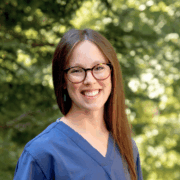 A woman with long straight brown hair is smiling at the camera. She is wearing glasses with dark round frames, turquoise earrings, and blue medical scrubs. The background is outdoors with green trees and soft sunlight