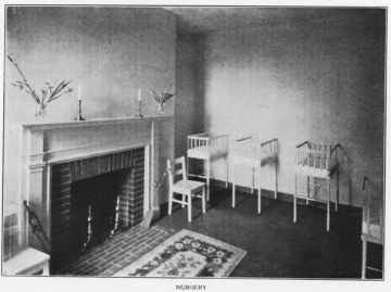 A black-and-white photograph of a nursery room with a fireplace, a chair, and several cribs along the wall. The room has a simple, yet cozy, appearance, reflecting the care environment for newborns in a historical hospital setting.