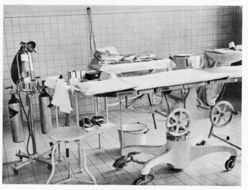 1923 - Operating room A black-and-white photograph of an early operating room with a surgical table, various medical instruments, oxygen tanks, and equipment. The room has tiled walls and a sterile appearance, highlighting the medical practices of the time.