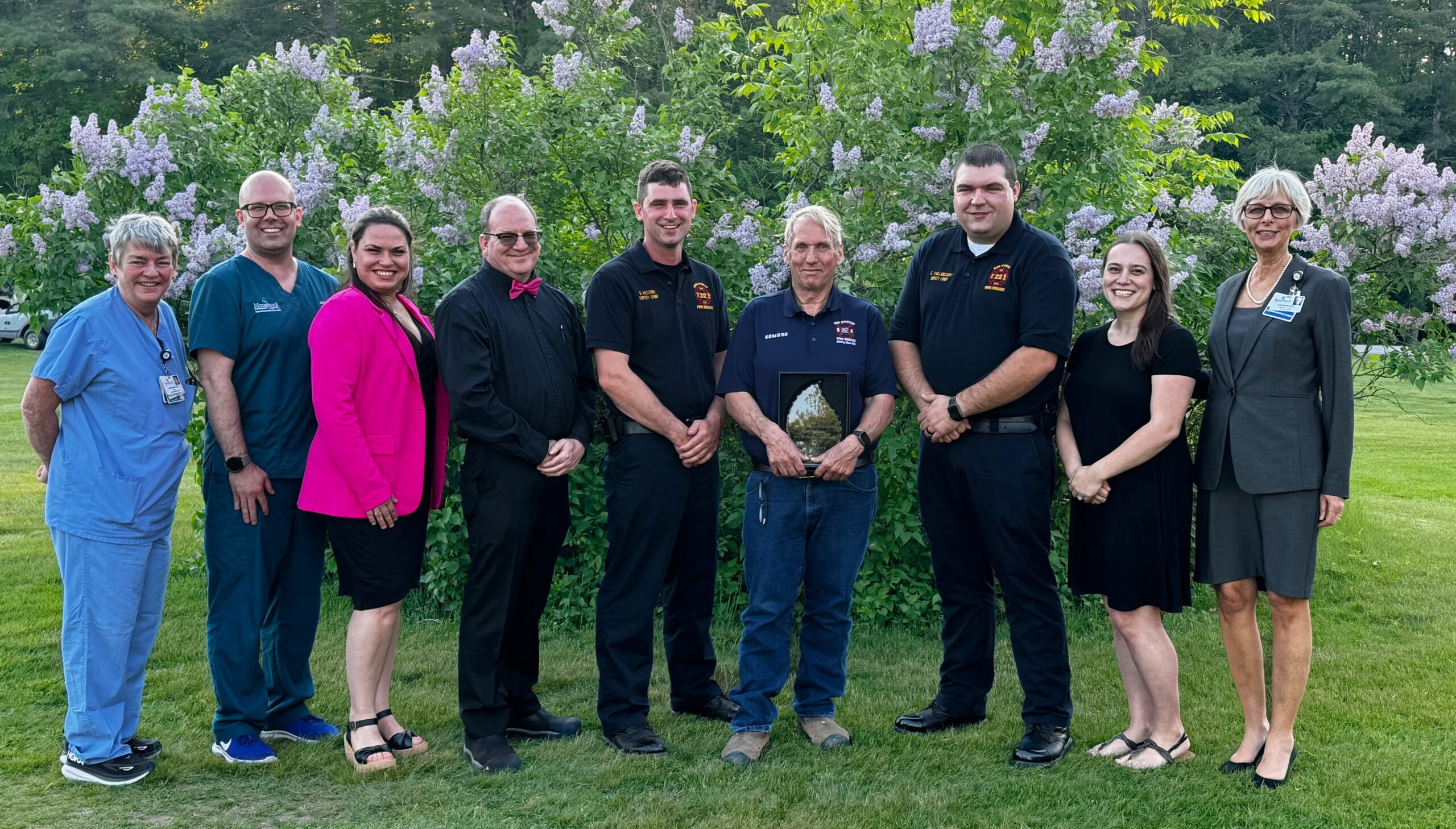 Lifetime Achievement recipient Gary Somero alongside members of the New Ipswich Fire Department and MCH leaders. A group of nine people, including EMS workers and hospital staff, standing in front of lilac bushes, smiling at the camera. They are being recognized for their service.